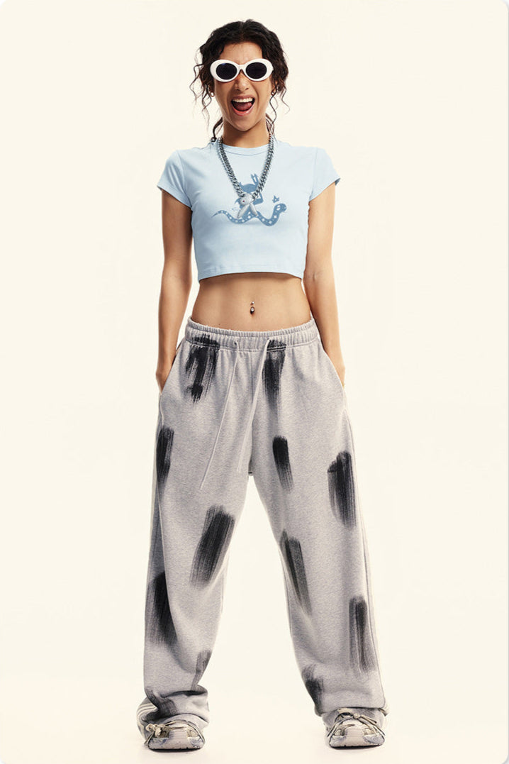 Model in a light blue graphic crop top and gray sweatpants, posing with sunglasses and a statement necklace