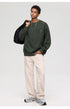 Male model holding black jacket, wearing olive textured sweatshirt and white pants with gray sneakers
