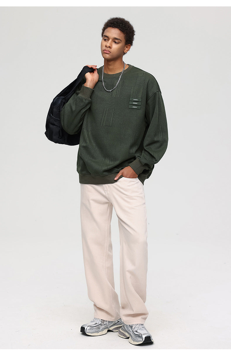 Male model holding black jacket, wearing olive textured sweatshirt and white pants with gray sneakers