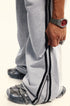 Close-up of ankle zipper detail on gray sweatpants with black double stripe trim, showing silver metal hardware and wristwatch