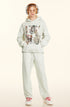 A woman is standing in a full-length pose wearing a light gray sweatsuit with graphic print hoodie, hands in pockets and red-accented sneakers