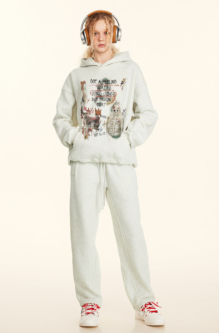 A woman is standing in a full-length pose wearing a light gray sweatsuit with graphic print hoodie, hands in pockets and red-accented sneakers