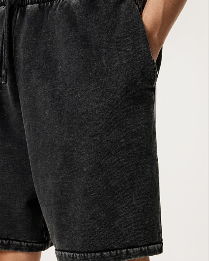 A close-up of black washed cotton shorts with elastic waistband showing pocket detail and hand reaching into side pocket