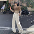 A woman is walking on a street holding ice cream, wearing a beige cropped sweater, white wide-leg pants and beige sneakers against a rustic brick wall