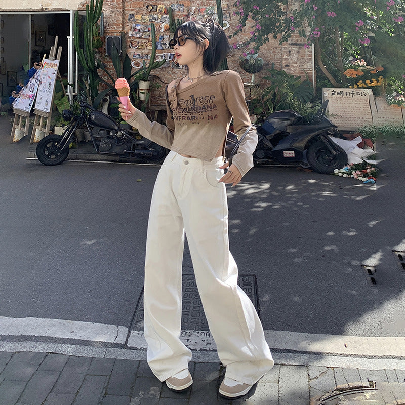 A woman is walking on a street holding ice cream, wearing a beige cropped sweater, white wide-leg pants and beige sneakers against a rustic brick wall