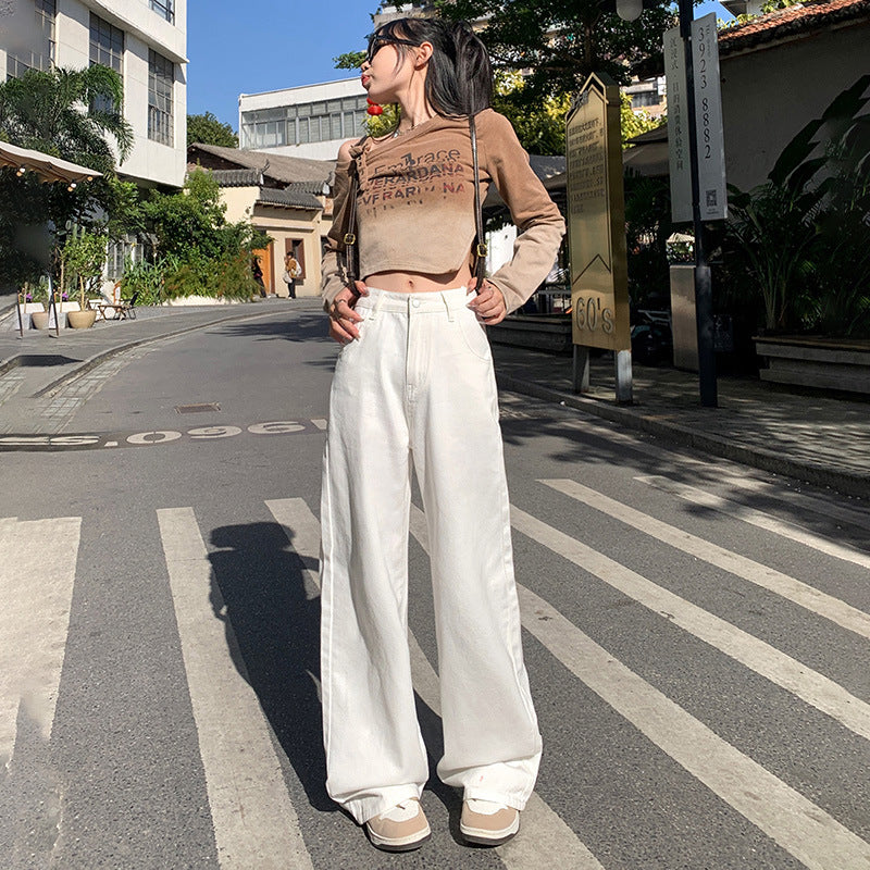 A woman is posing on a crosswalk wearing a beige cropped sweater with text, white high-waisted wide-leg pants and beige sneakers