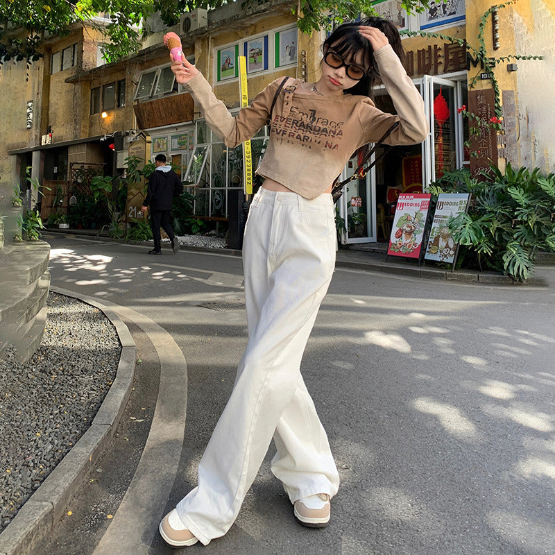 A woman is standing with ice cream, touching her hair, wearing a beige cropped sweater, white wide-leg pants and sneakers outside a storefront with yellow walls and greenery