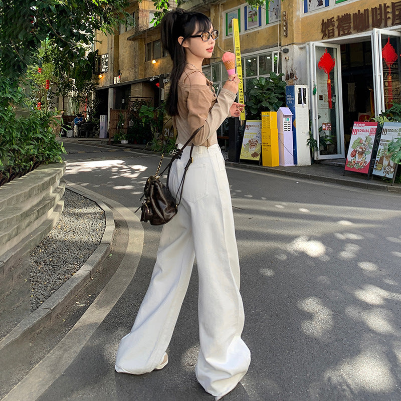 A woman is standing sideways holding ice cream, wearing a beige cropped top, white wide-leg pants, black glasses and crossbody bag on a tree-lined street with shops