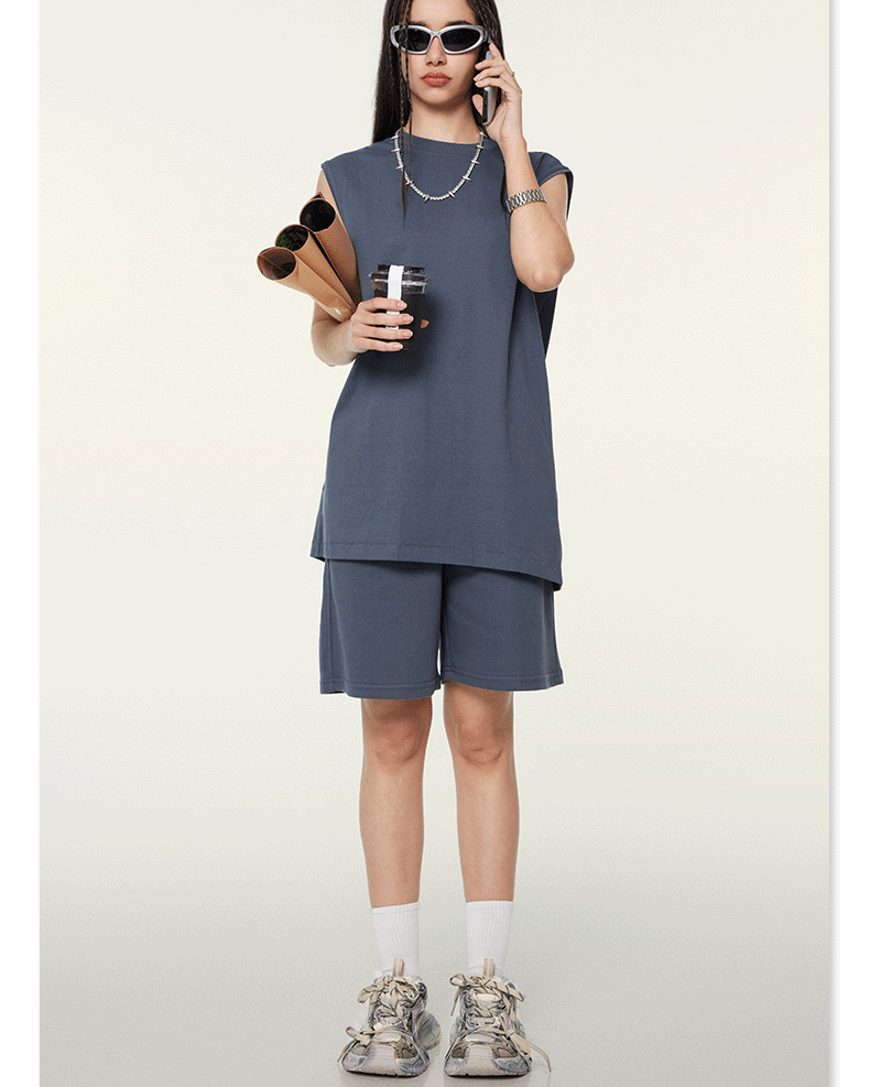 Model wearing slate gray matching set with sleeveless top and shorts, talking on phone while holding documents, wearing sunglasses and silver chain necklace