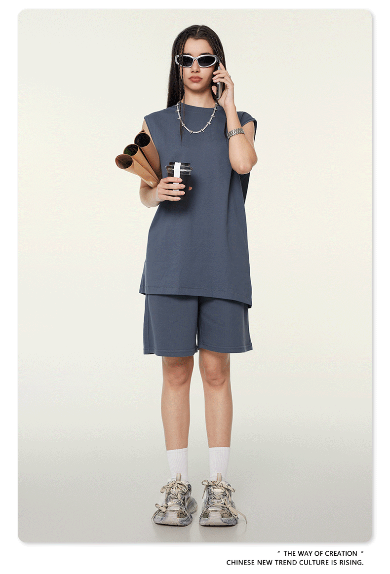 Model wearing slate gray matching set with sleeveless top and shorts, talking on phone while holding documents, wearing sunglasses and silver chain necklace