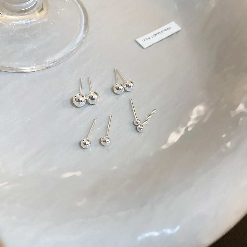 A pair of silver-tone ball stud earrings in various sizes, arranged on a dish with a reflective surface.