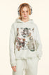A woman is wearing headphones while modeling a light gray graphic hoodie with artistic prints and text, featuring front pocket detail