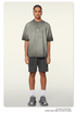 Model with braided hair wearing gray ombre t-shirt with silver pendant necklace, dark cargo shorts and white socks against white background