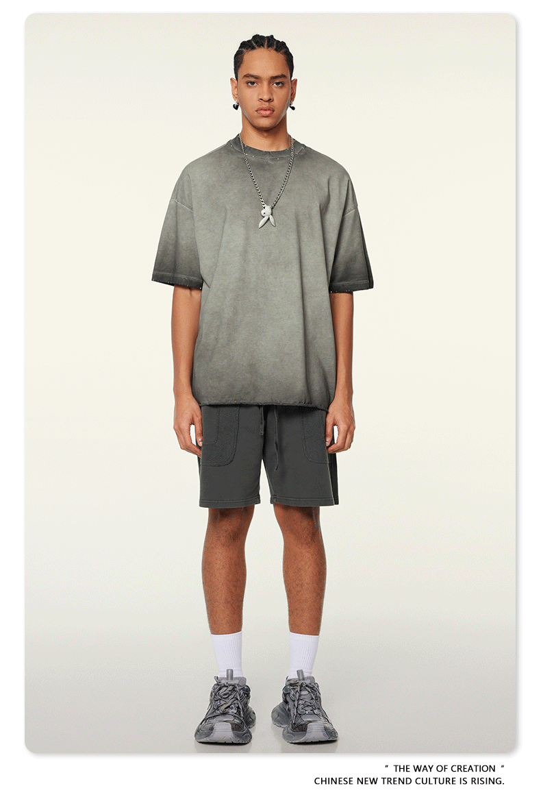Model with braided hair wearing gray ombre t-shirt with silver pendant necklace, dark cargo shorts and white socks against white background