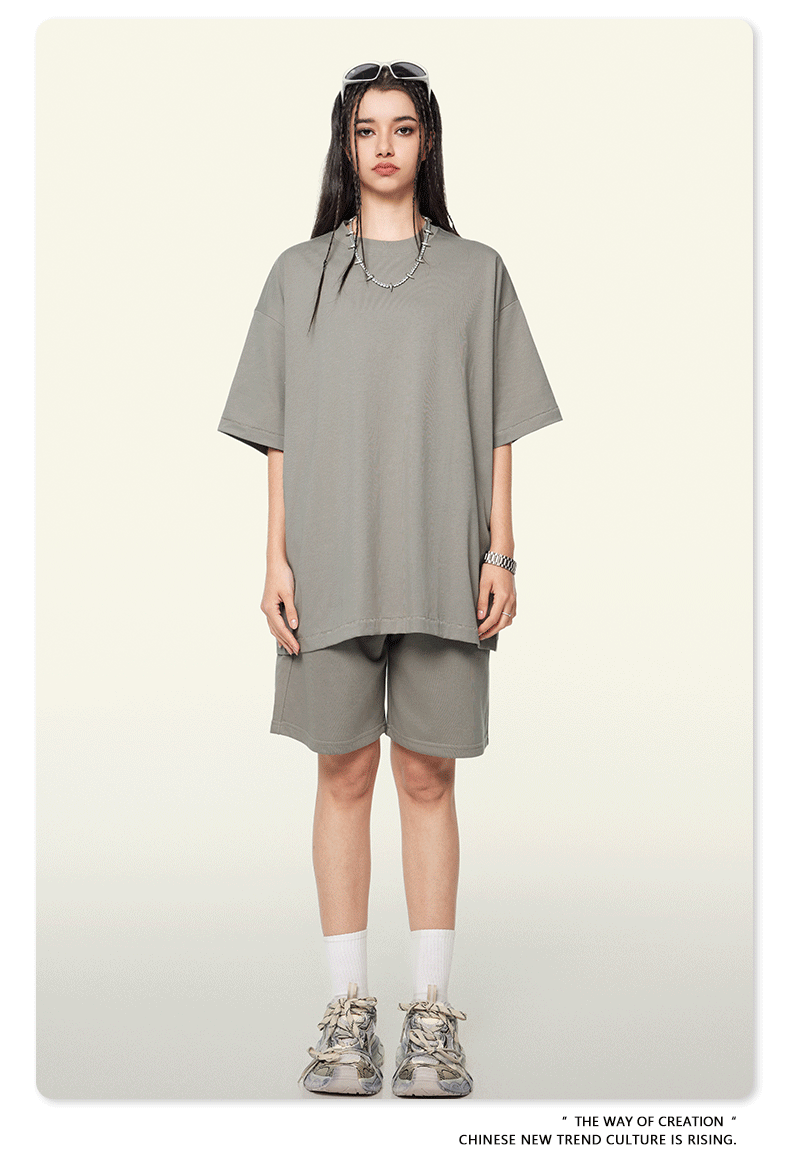 A woman wearing a gray oversized t-shirt and matching shorts set, accessorized with sunglasses on head, silver necklace, and white sneakers against a neutral backdrop