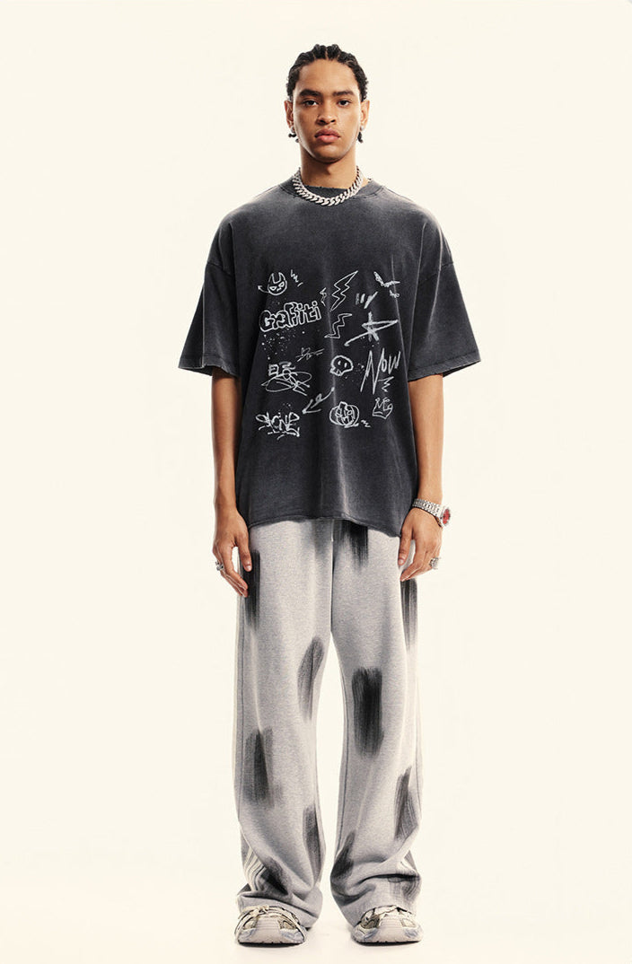 A model wearing black graffiti-print oversized t-shirt and light gray tie-dye pants against white backdrop, silver chain necklace