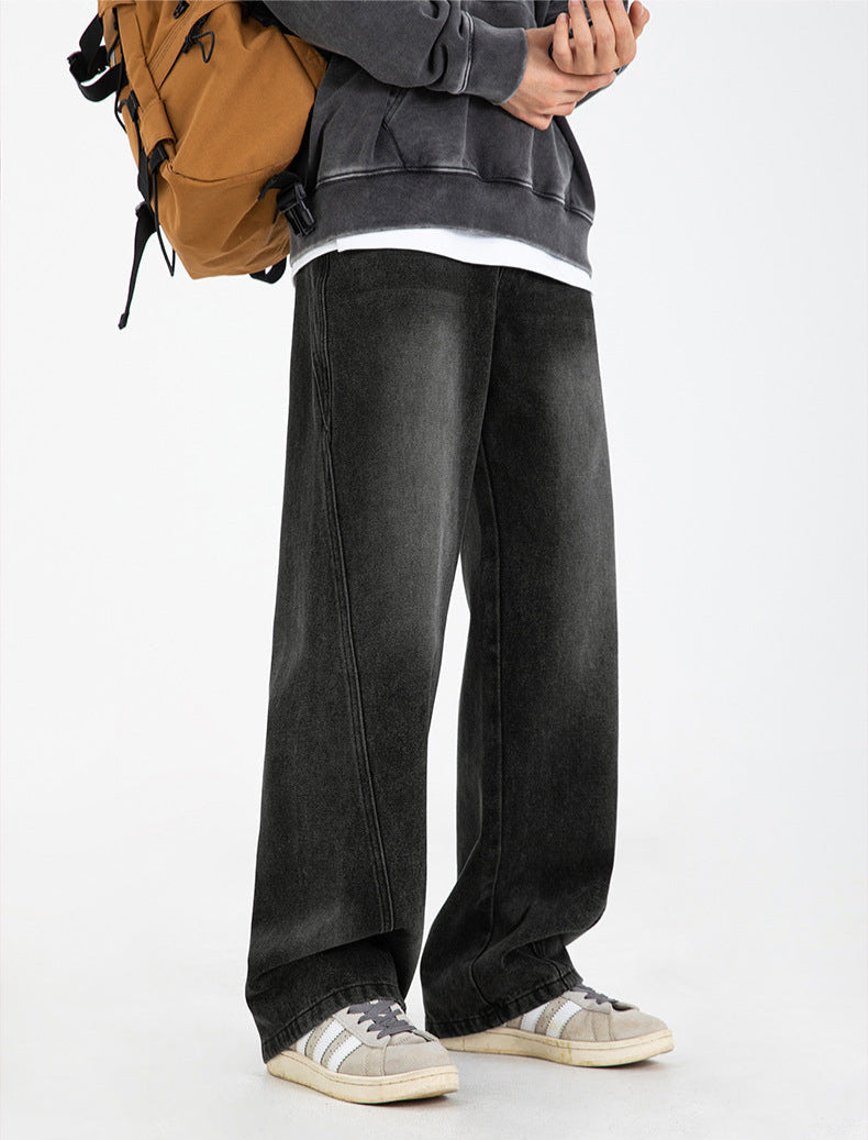 A model standing wearing black wide-leg jeans with distinctive seams, gray sweatshirt layered over white tee, tan backpack, and retro gray sneakers with white stripes