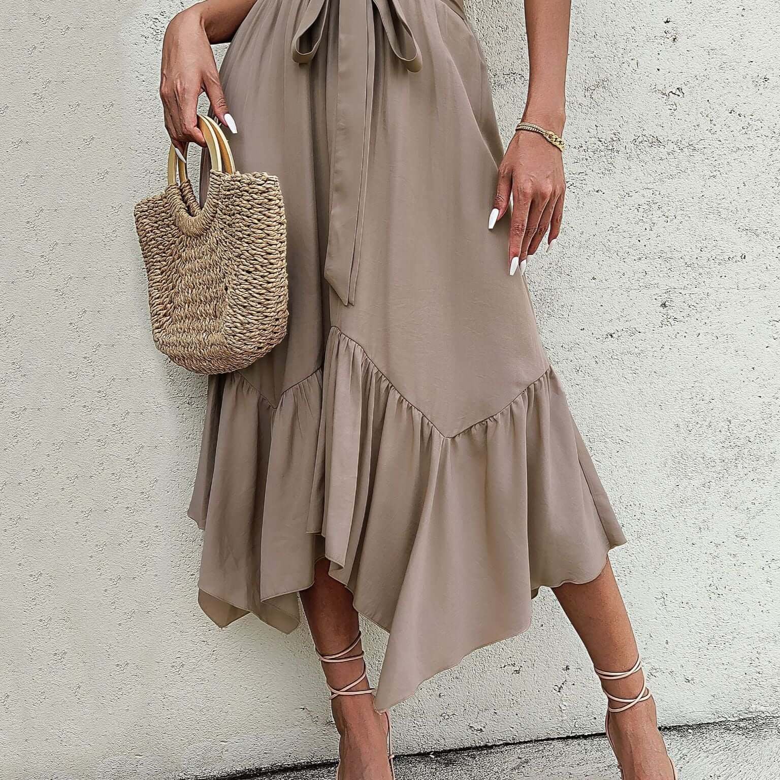 Woman wearing a khaki asymmetrical ruffle midi skirt with a tie waist, styled with strappy heels and a woven bag.