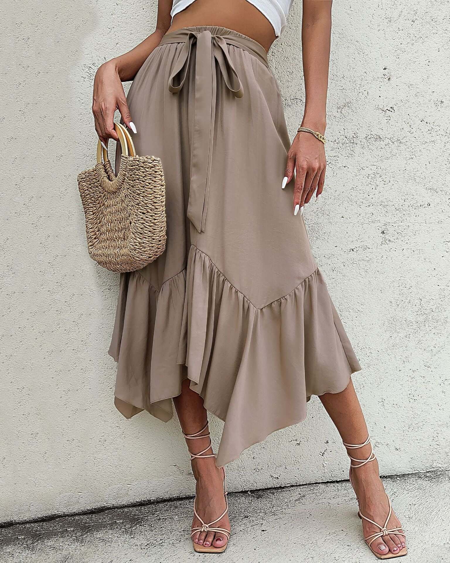 Woman wearing a khaki asymmetrical ruffle midi skirt with a tie waist, styled with strappy heels and a woven bag.