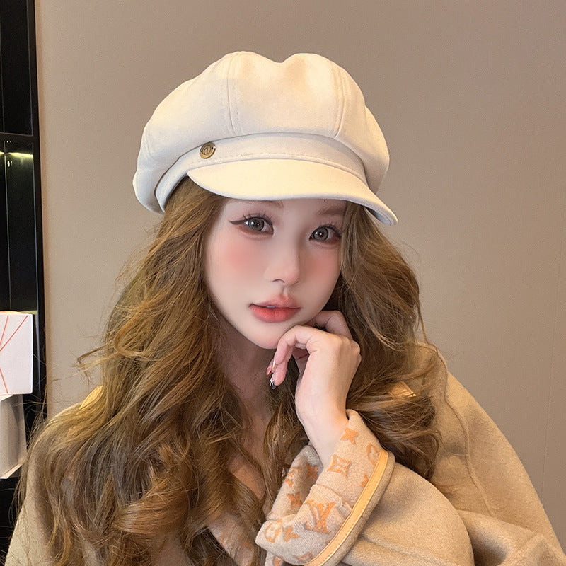 Woman wearing white newsboy cap with small metal emblem and wavy brown hair against neutral background