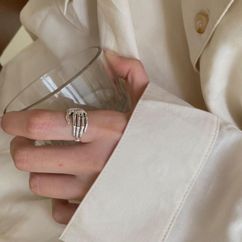 Close-up of a silver skeleton hand ring on a person's finger, holding a glass, styled with a cream-colored shirt