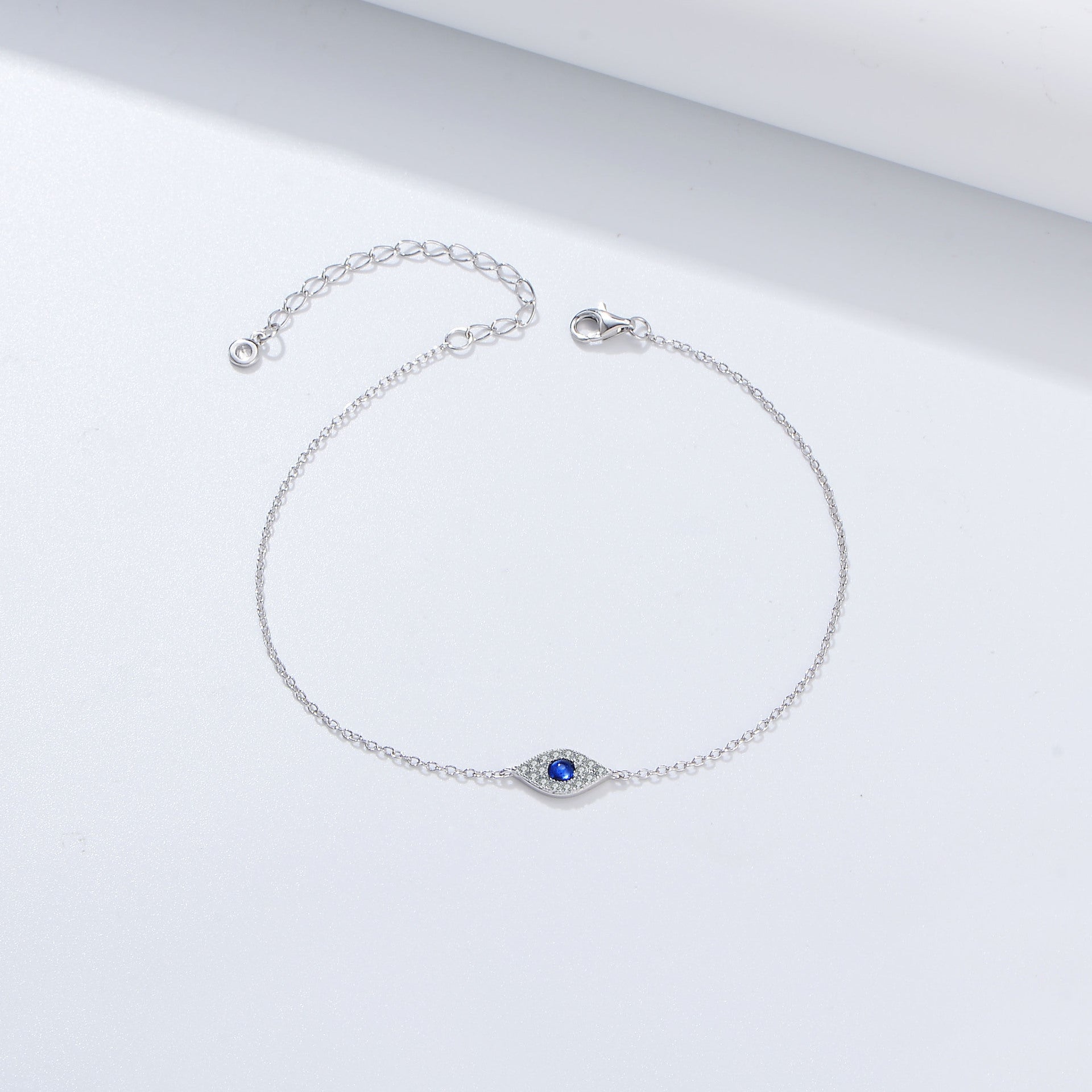 Silver bracelet with evil eye charm featuring blue sapphire center and diamond pavé halo, displayed on white surface with extension chain