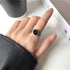 A woman wearing a silver timepiece ring with black dial and watch hands on ring finger against white desktop with black leather accessory