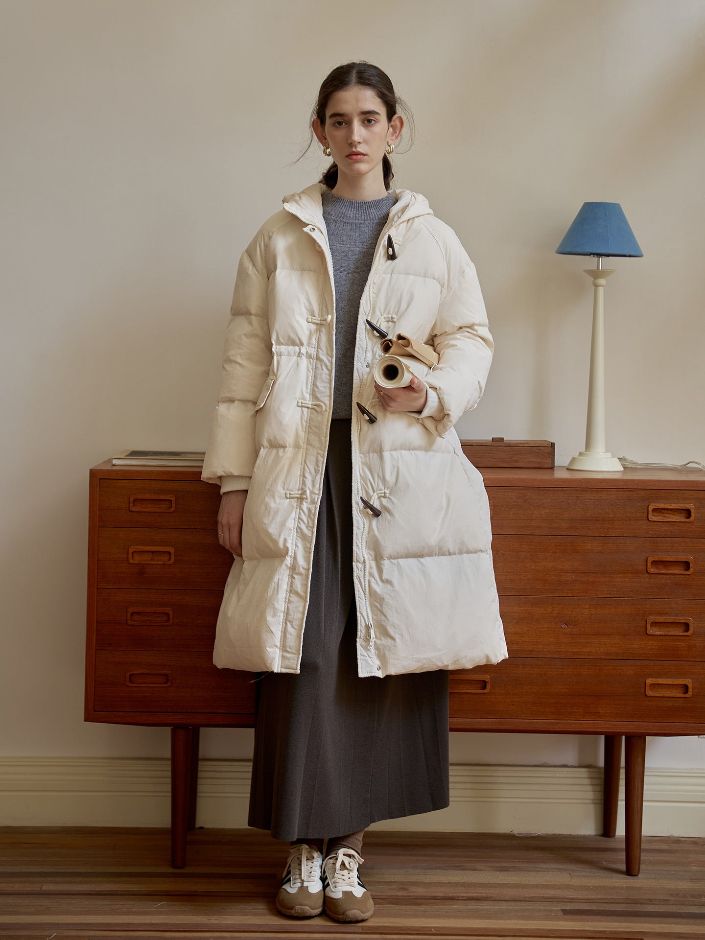 Long white quilted puffer coat with toggle closures over gray sweater and skirt, styled with vintage dresser and blue lamp