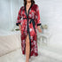 Red floral satin robe with black trim, tied at waist, worn open to reveal leg, displayed on white textured background