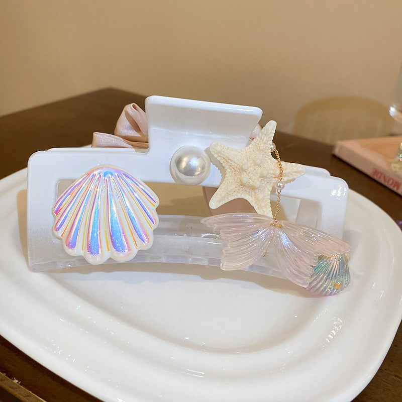 A white hair claw clip decorated with a pearl, a textured starfish charm, an iridescent seashell, and a dangling translucent mermaid fin charm.