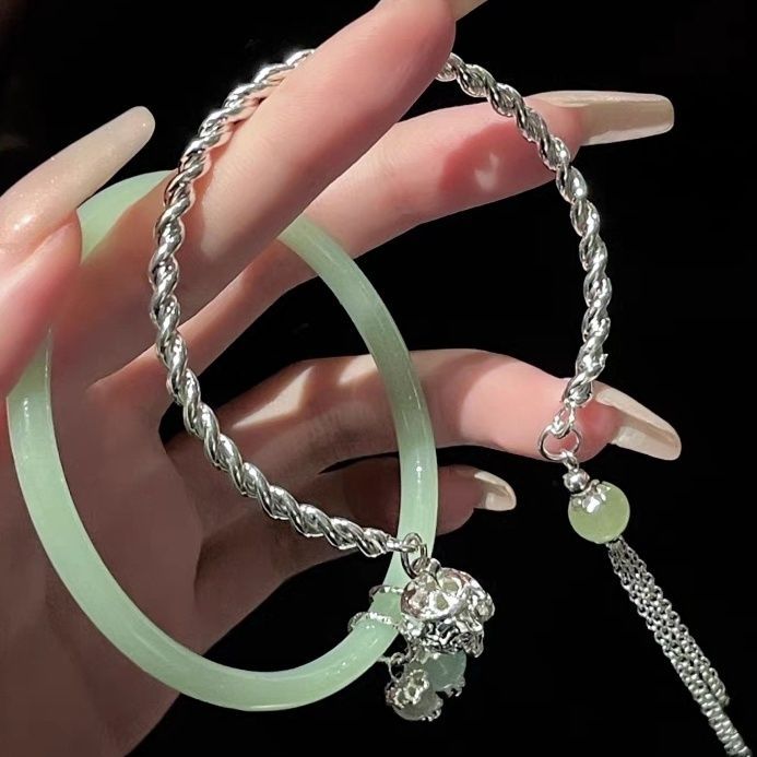 A woman holding a silver twisted bracelet and light jade bangle with decorative charms against black background