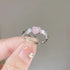 A person holding a silver ring with pink heart-shaped gemstone center flanked by clear crystals, showing decorated fingernails against neutral background