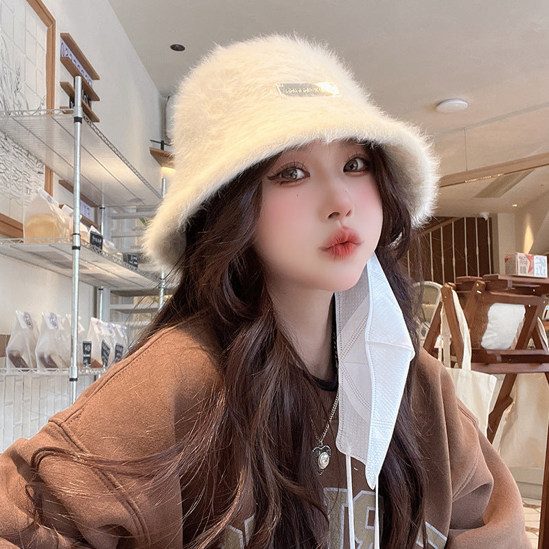 Woman wearing a white fluffy bucket hat in a cozy café setting, dressed in a brown sweater with a mask hanging from her ear