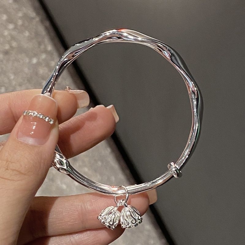 A person is holding a silver twisted bangle bracelet with decorative bell-shaped charms, featuring a French manicure with rhinestone nail art against a dark gray background