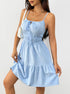 A woman is wearing a light blue spaghetti strap sundress with shirred bodice and ruffle hem, carrying a white handbag against a white background