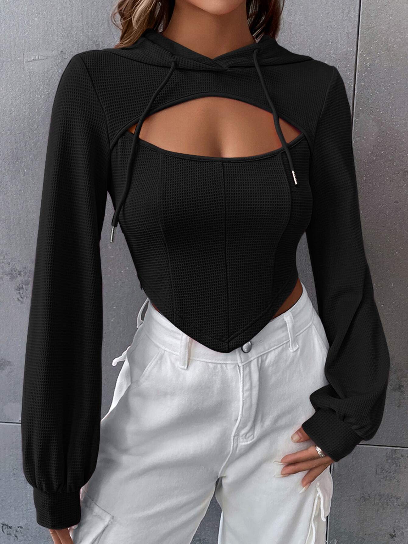 Woman wearing black textured crop hoodie with chest cutout detail paired with white cargo pants against gray wall