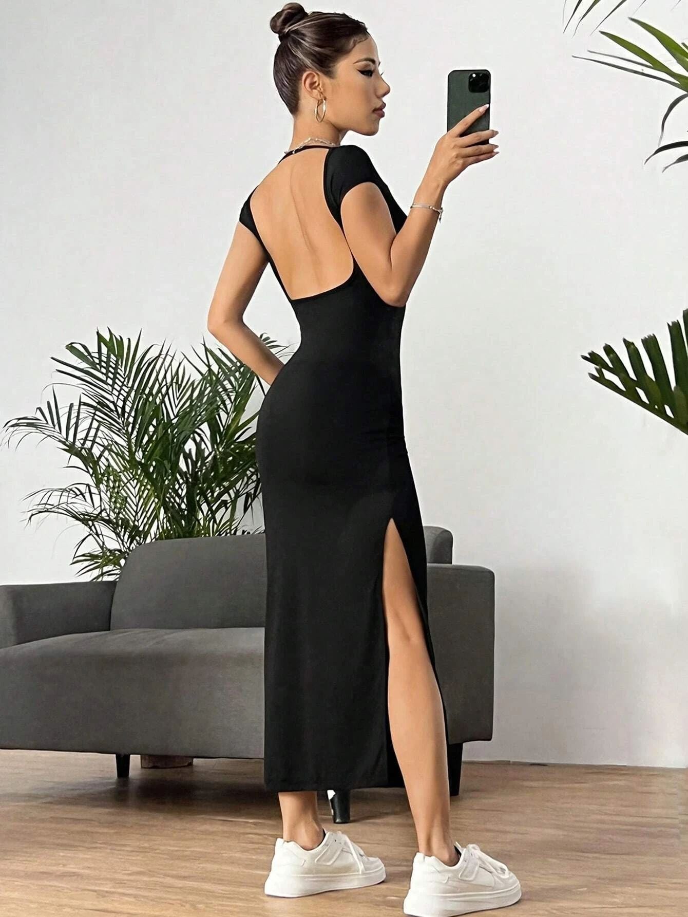 A woman taking a mirror selfie in a black maxi dress with open back, side slit, and cap sleeves, styled with white sneakers