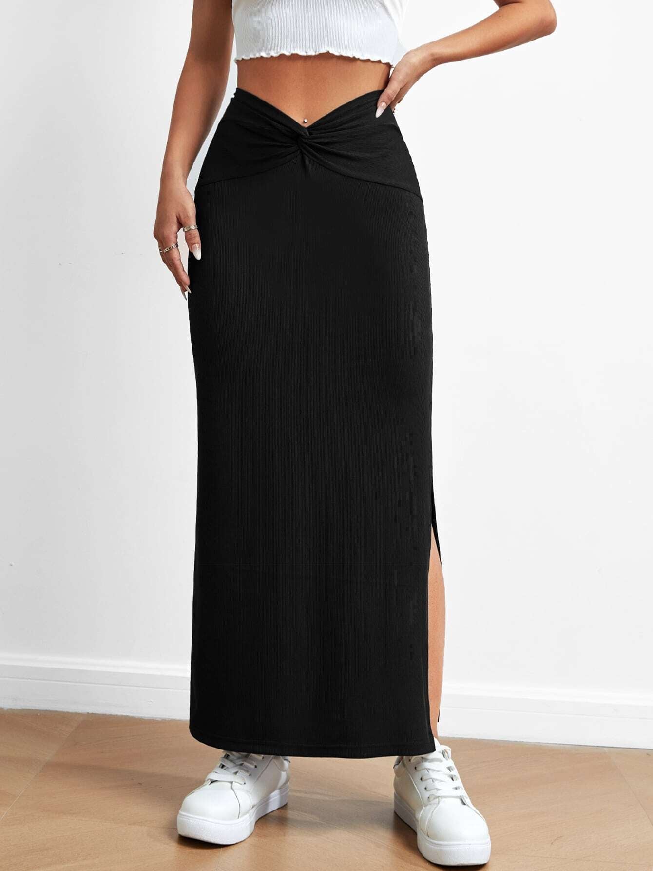 Black long skirt with side slit and twisted waistband, paired with white sneakers and a white cropped top, photographed against a light background