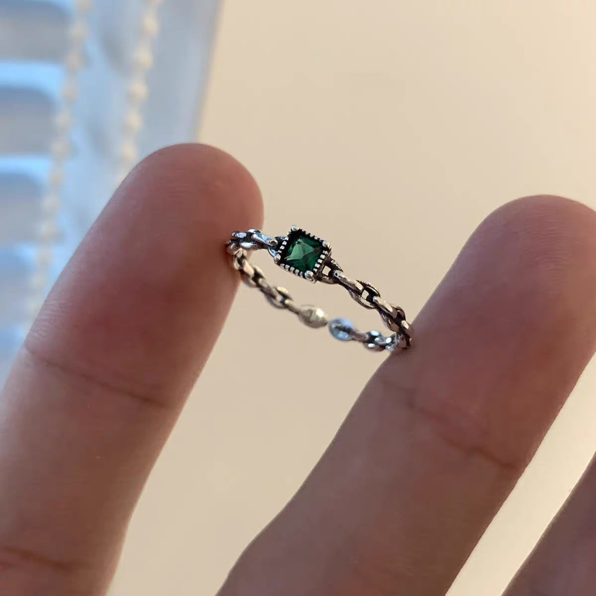 A delicate silver chain ring with square emerald green gemstone centered between fingers against neutral background