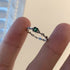 A delicate silver chain ring with square emerald green gemstone centered between fingers against neutral background