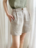 A model wearing beige linen shorts with pleated front detail and high waist, paired with a sage green top against sheer curtains