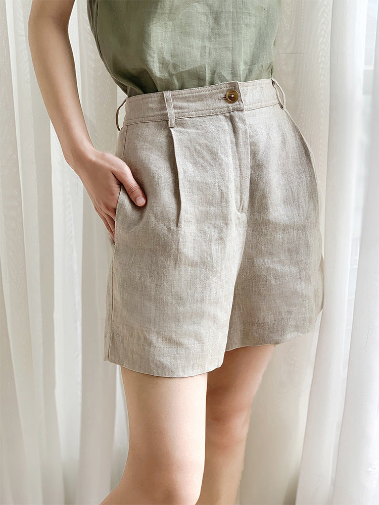 A model wearing beige linen shorts with pleated front detail and high waist, paired with a sage green top against sheer curtains