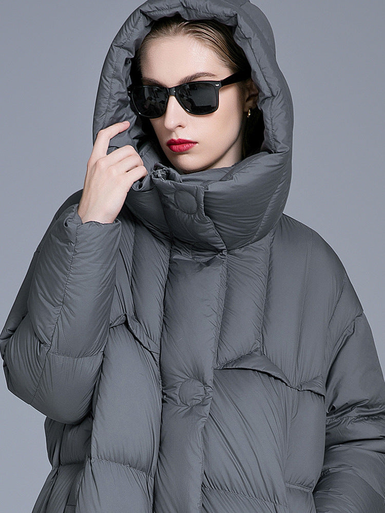 A woman wearing a gray oversized puffer coat with hood pulled up, black sunglasses and red lipstick, touching hood while posing against gray background