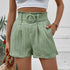 Model wearing sage green pleated shorts with matching belt, white crop top, and carrying woven bag against white wall backdrop
