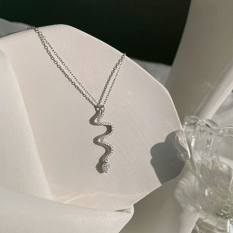 A silver snake pendant necklace with crystal accents displayed on white fabric with glass decor nearby