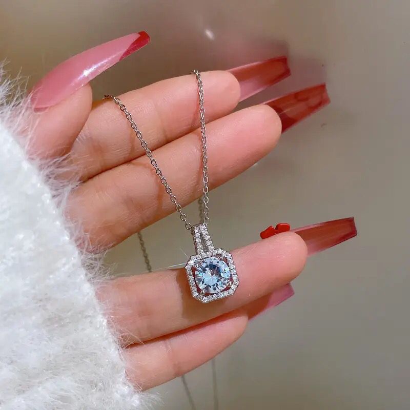 Blue crystal square halo necklace with cubic zirconia accents shown on hand with red manicure, wearing white sweater