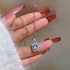 Blue crystal square halo necklace with cubic zirconia accents shown on hand with red manicure, wearing white sweater