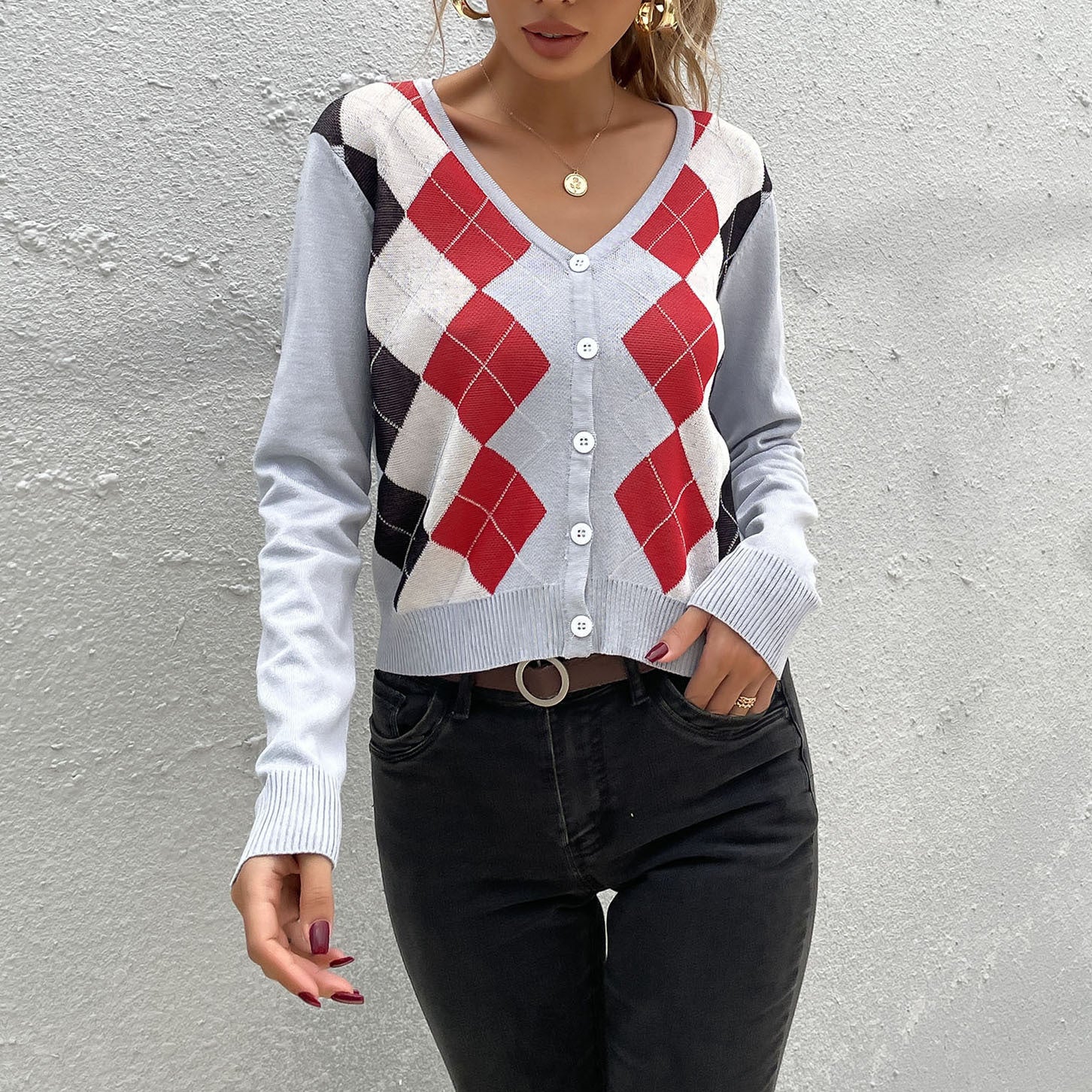 A woman wearing a light gray argyle cardigan with red and black pattern, paired with black jeans against textured white wall