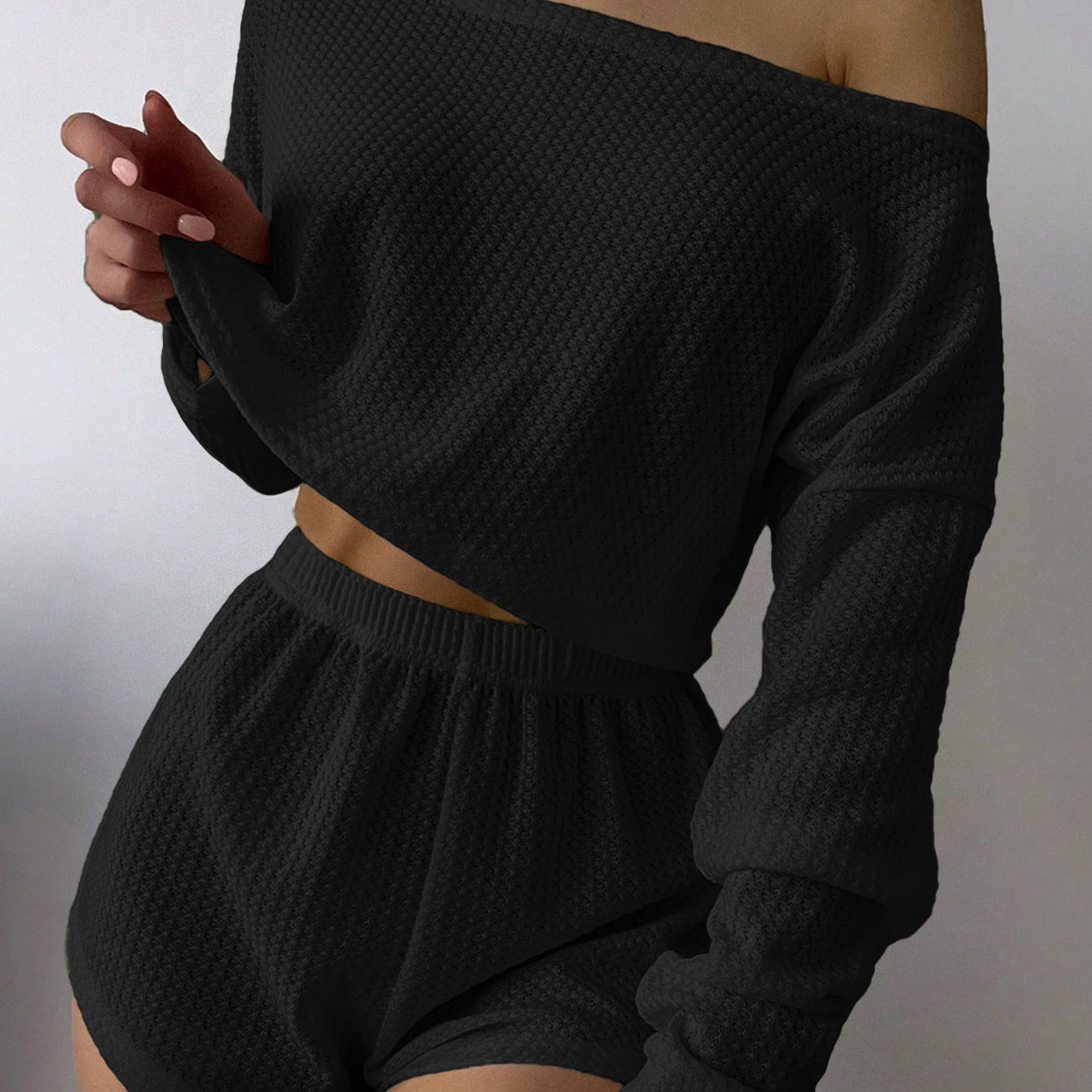 Woman wearing black off-shoulder knit two-piece loungewear set with crop top and shorts against white background