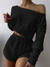 Woman wearing black off-shoulder knit two-piece loungewear set with crop top and shorts against white background